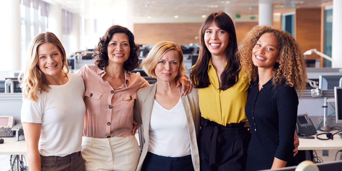 Five women in an office standing with their arms around each other.